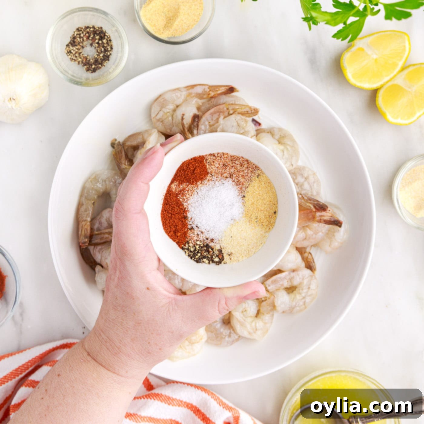 Sprinkling a bowl of mixed spices over butter-coated raw shrimp in a mixing bowl.