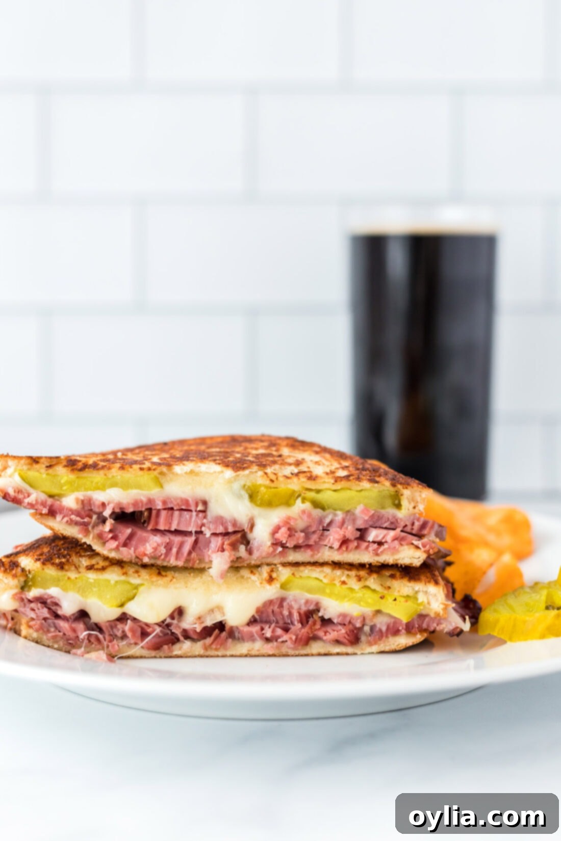 Delicious grilled corned beef sandwich on a white plate, cut in half to showcase the succulent beef, melted mozzarella, and crisp pickle layers.