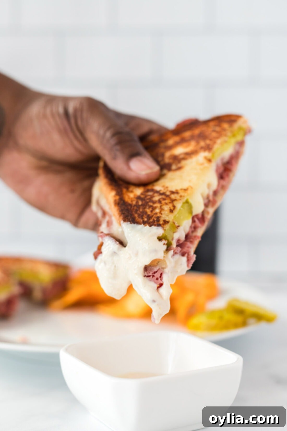 A hand dipping a grilled corned beef sandwich into a small ramekin of horseradish mayo, highlighting the golden-brown crust.