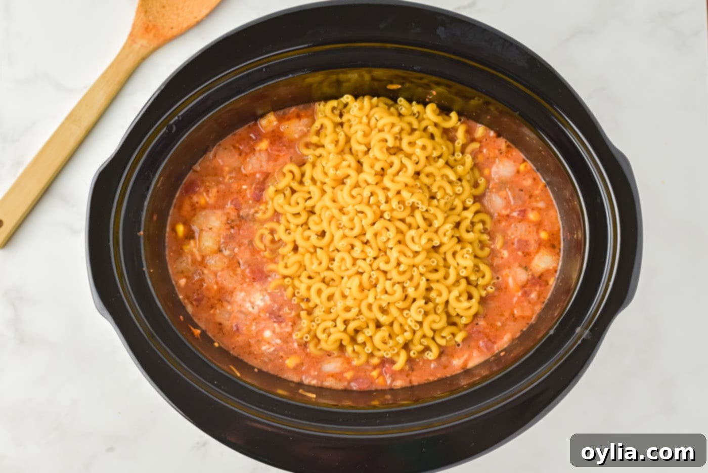 uncooked macaroni pasta on top of ground beef casserole in a slow cooker, ready to be stirred in