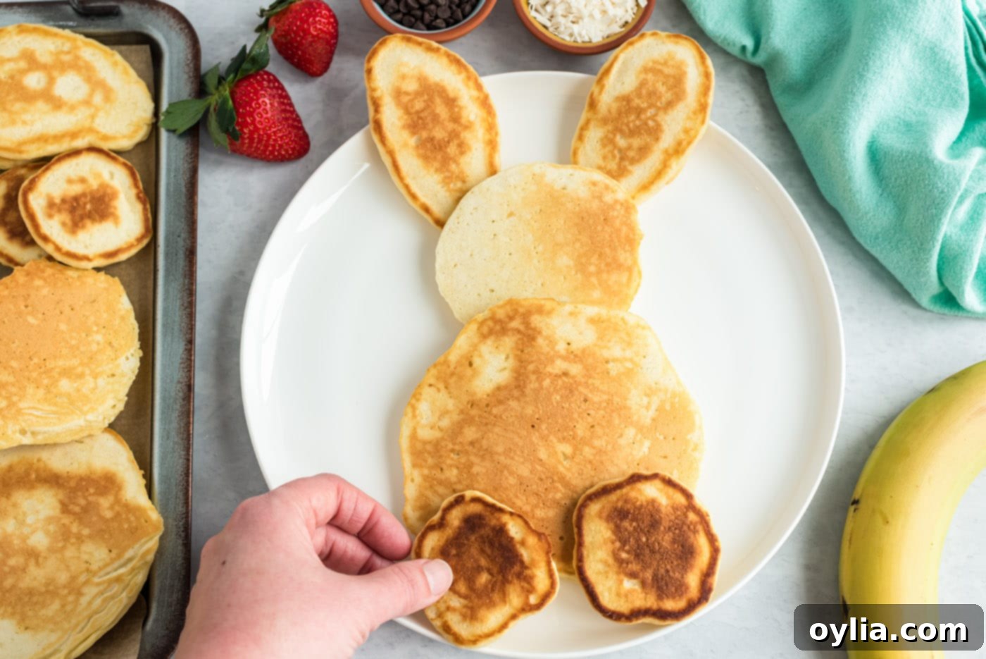A completed bunny butt pancake on a plate, featuring a large pancake body, smaller head, oval ears, banana slice feet with blueberry toes, and a whipped cream tail.