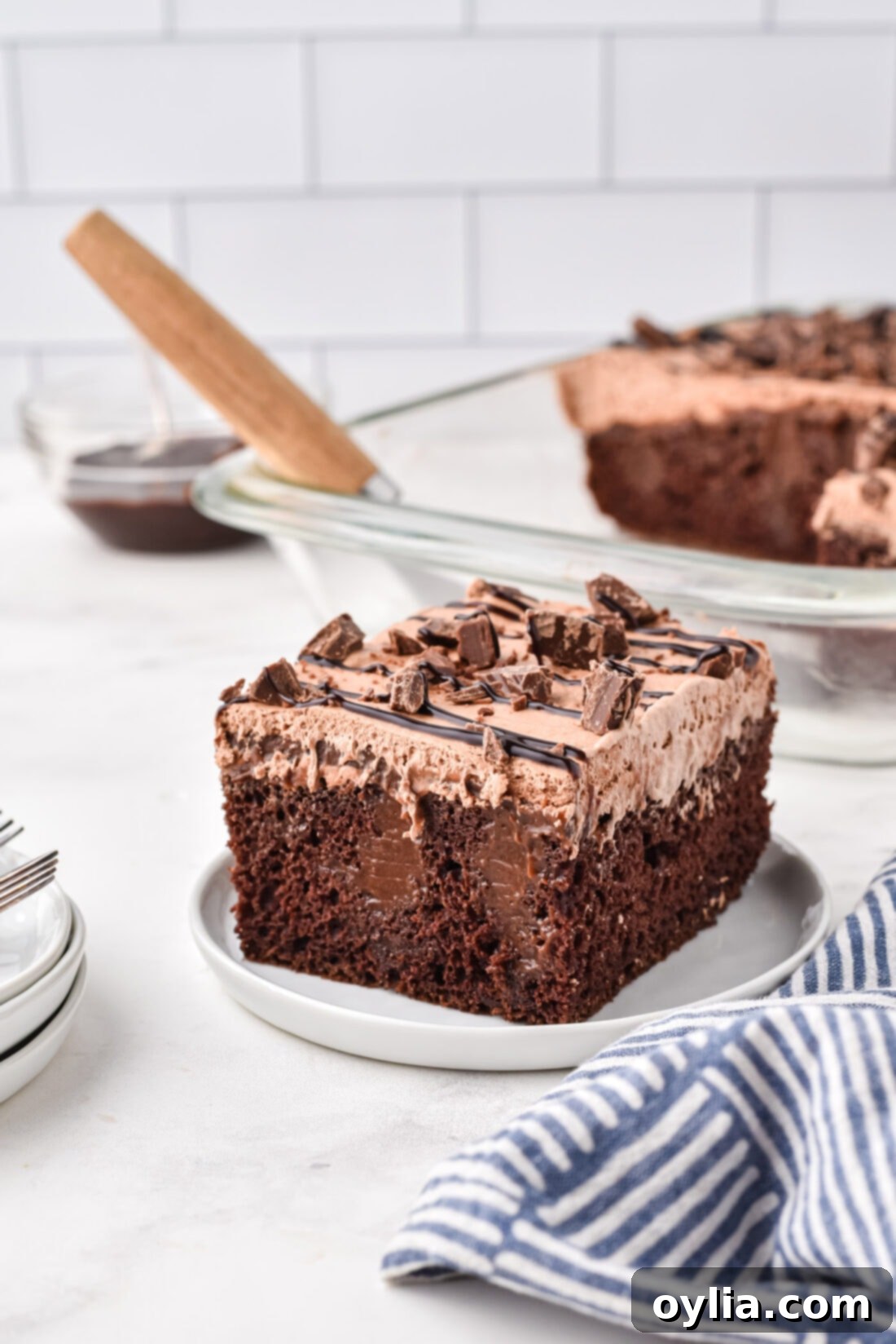 Decadent Chocolate Poke Cake 2 slice of incredibly moist and decadent Triple Chocolate Poke Cake on a white plate, showcasing its layers of cake, pudding, and whipped topping, garnished with chocolate drizzle and chopped candy.