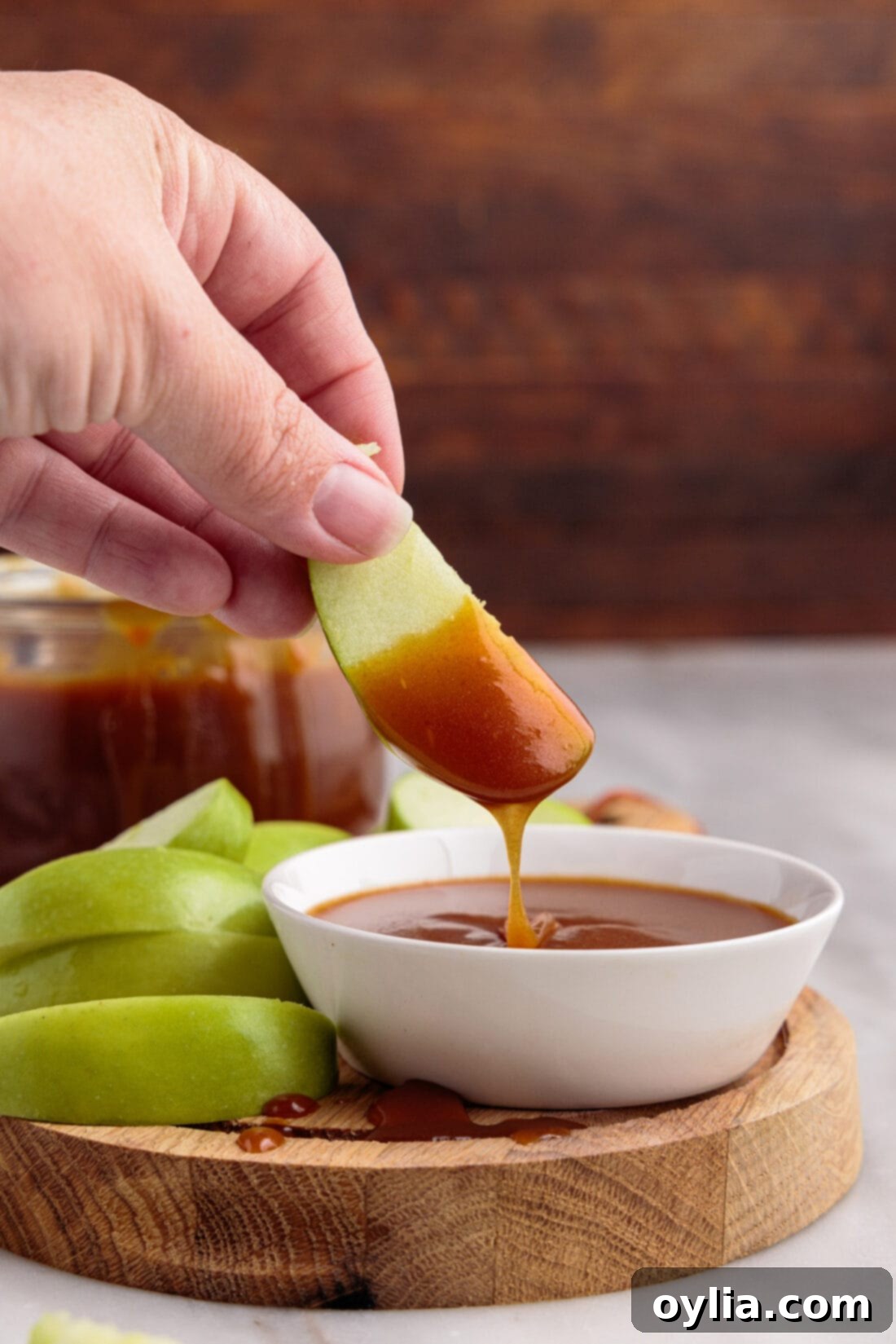 Apple slice being dipped into a jar of creamy, amber-colored Homemade Caramel Sauce.