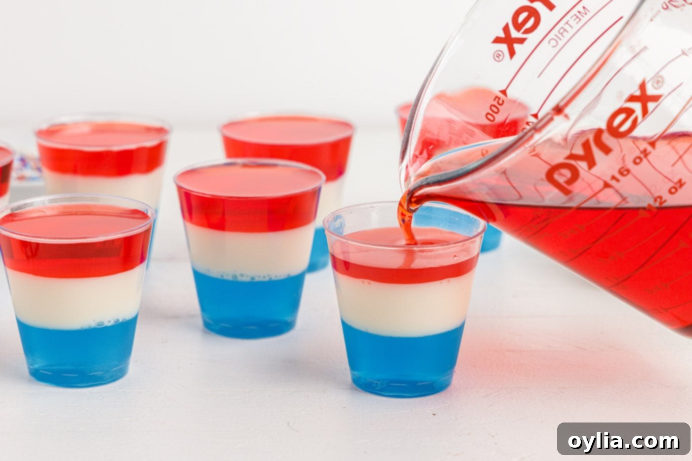 Pouring the final red jello layer over the set white and blue layers in shot glasses