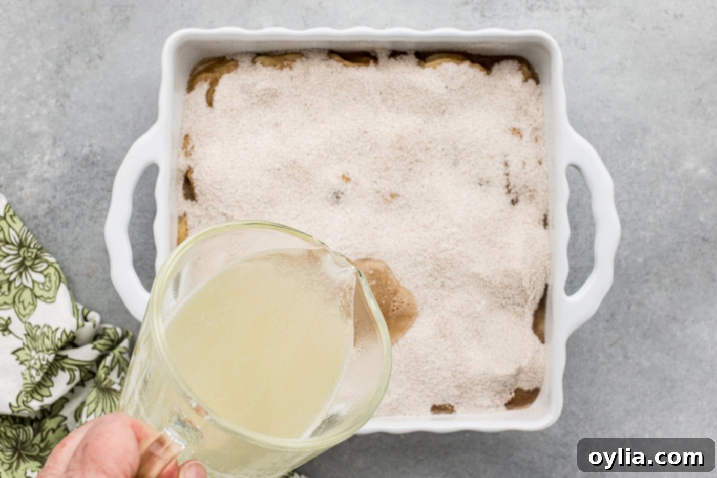 The process of pouring heated, boiling fruit juices from a glass measuring cup over the top of the unbaked cobbler with its spiced sugar layer.