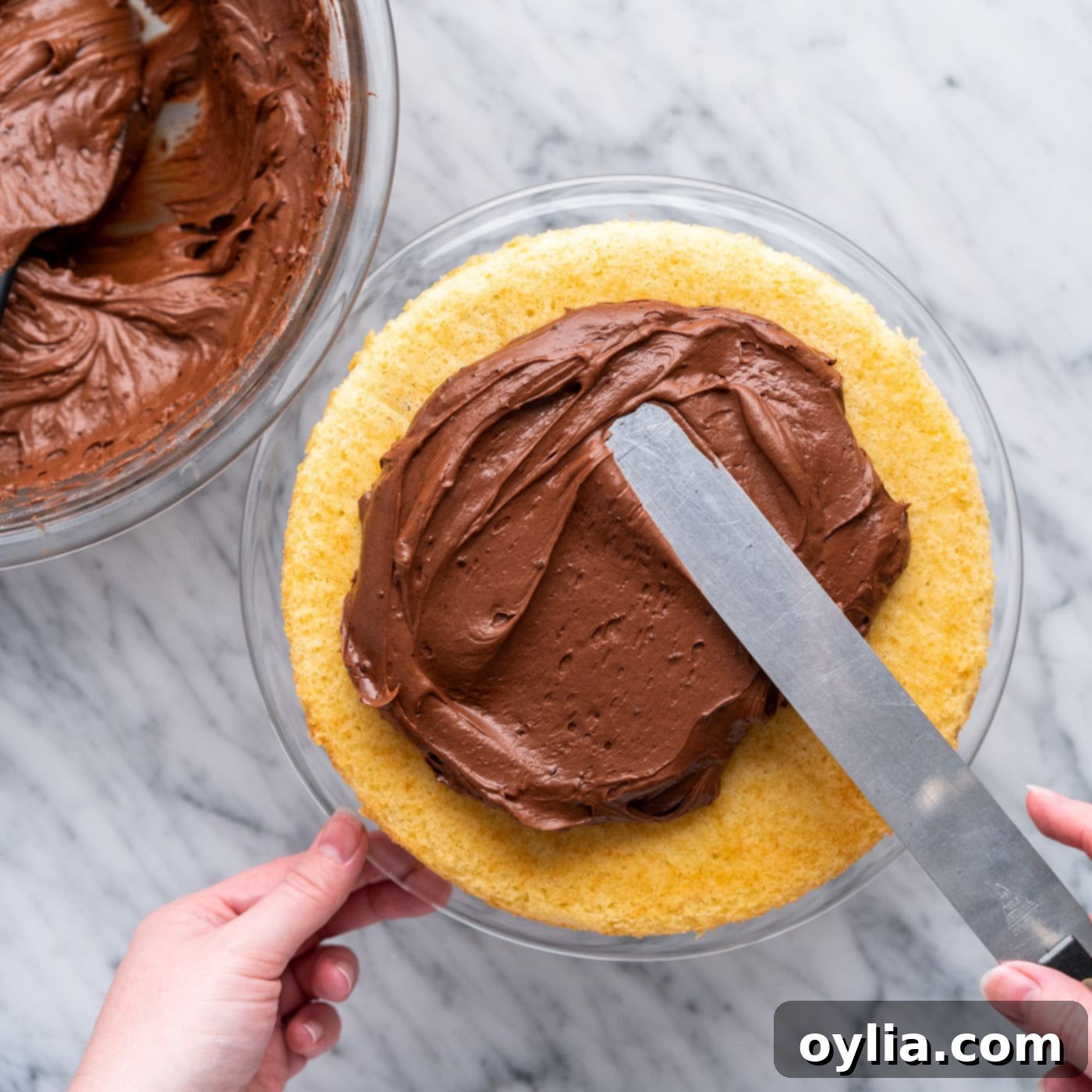 First layer of marble cake on a platter, being frosted with chocolate fudge frosting