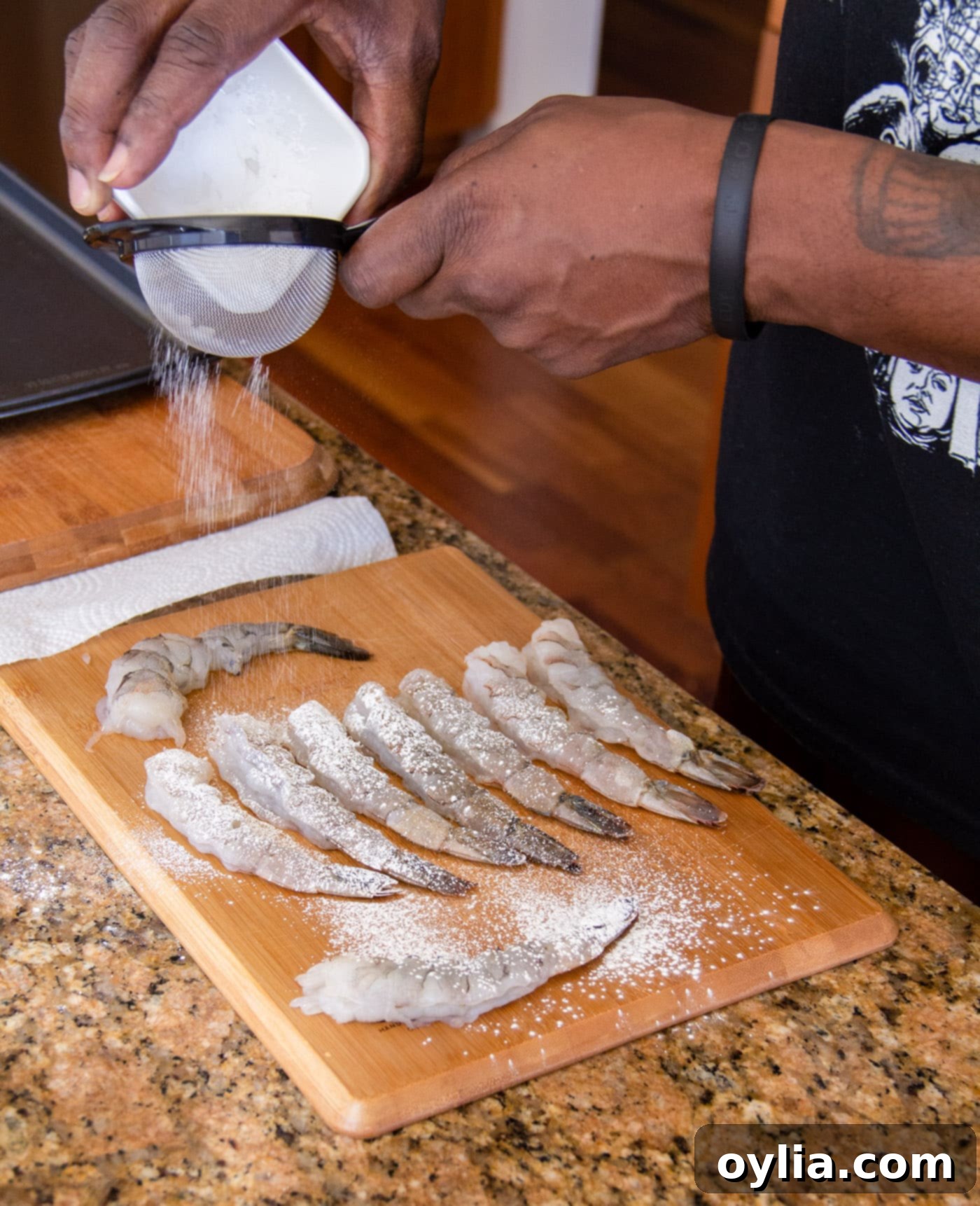 Sifting a fine layer of cornstarch over raw shrimp on a cutting board, preparing them for batter.