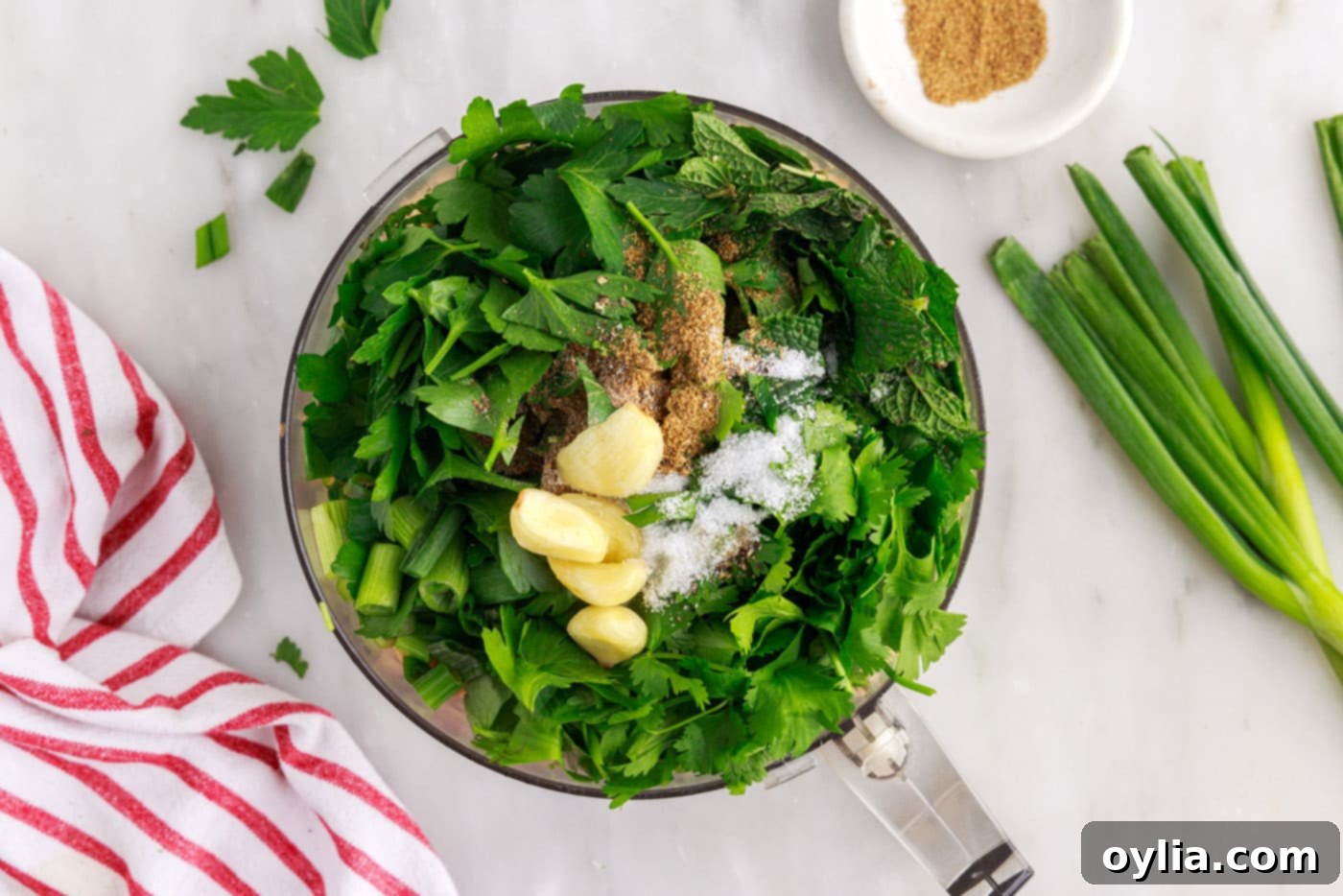Fresh herbs and garlic being added to a food processor containing soaked chickpeas.