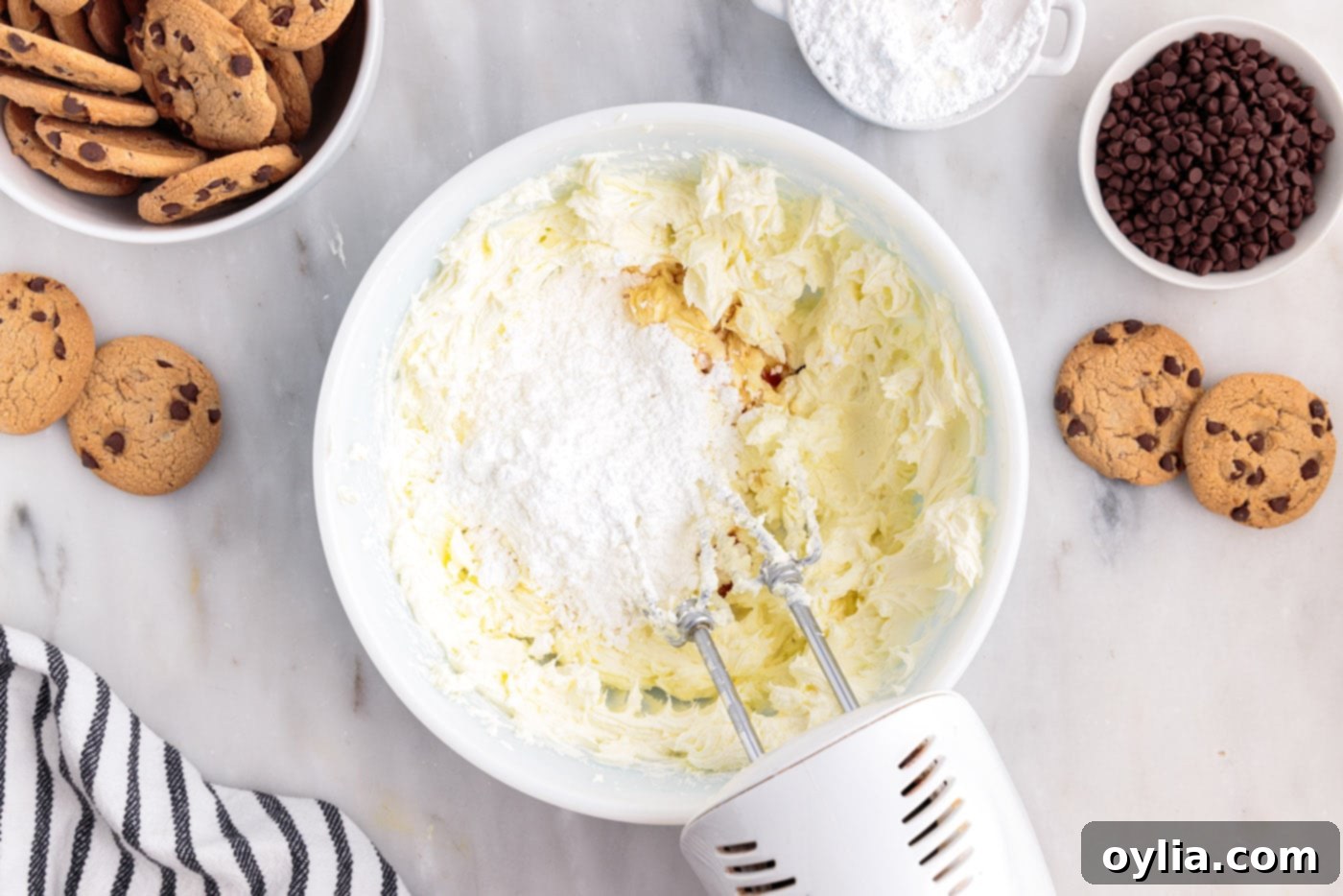 Powdered sugar and vanilla extract being added to softened cream cheese in a mixing bowl and beaten until smooth