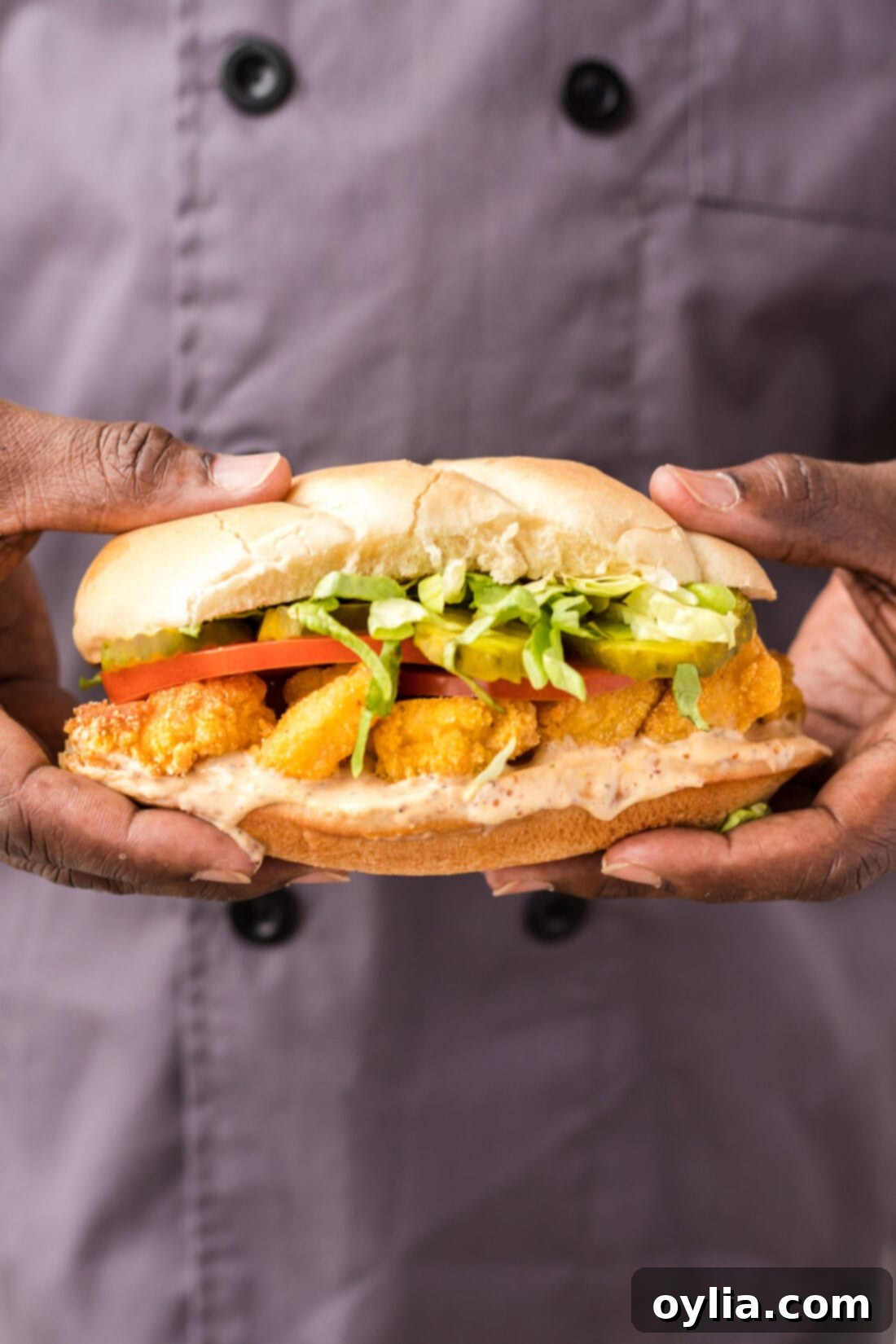 Chef holding a perfectly assembled Crispy Shrimp Po Boy sandwich, showcasing the golden-fried shrimp and fresh toppings.