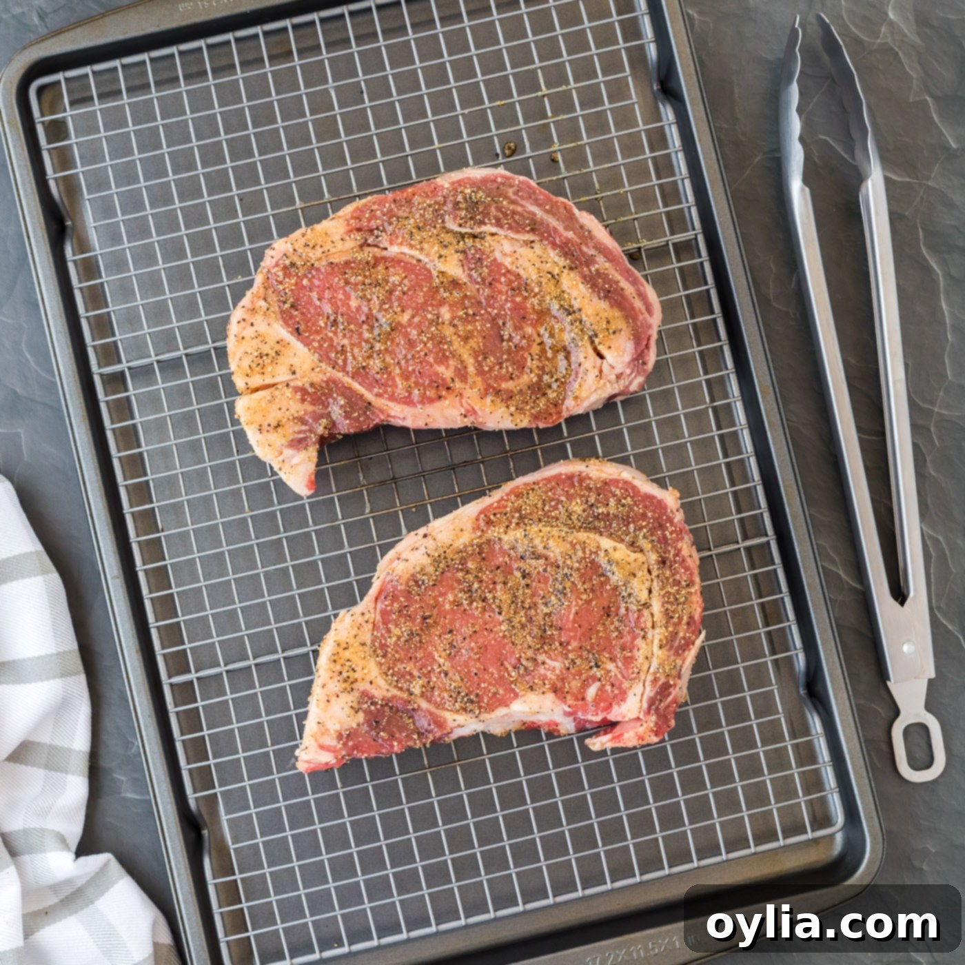 Raw ribeye steak, thoroughly rubbed with spices and olive oil, resting on a wire rack over a baking sheet.