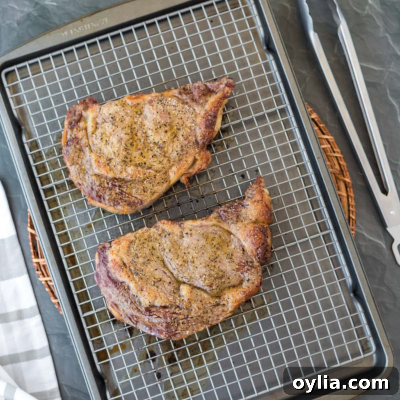 Two baked ribeye steaks resting on a wire rack over a baking sheet, just removed from the oven and before searing.