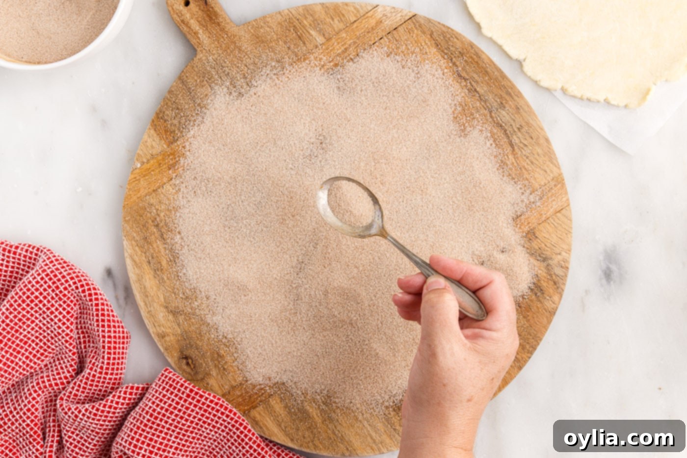A hand sprinkling cinnamon sugar evenly over a cutting board, creating a base for the rugelach dough.