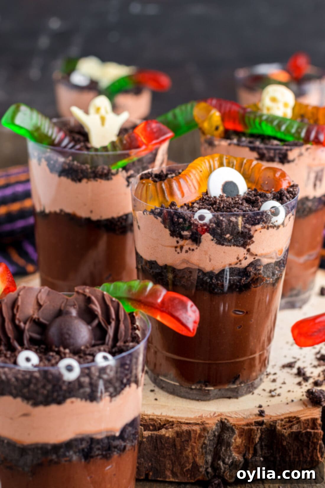 Two Halloween Dirt Cups on a rustic wood board, one with gummy worms, the other with candy eyeballs and a chocolate pumpkin.