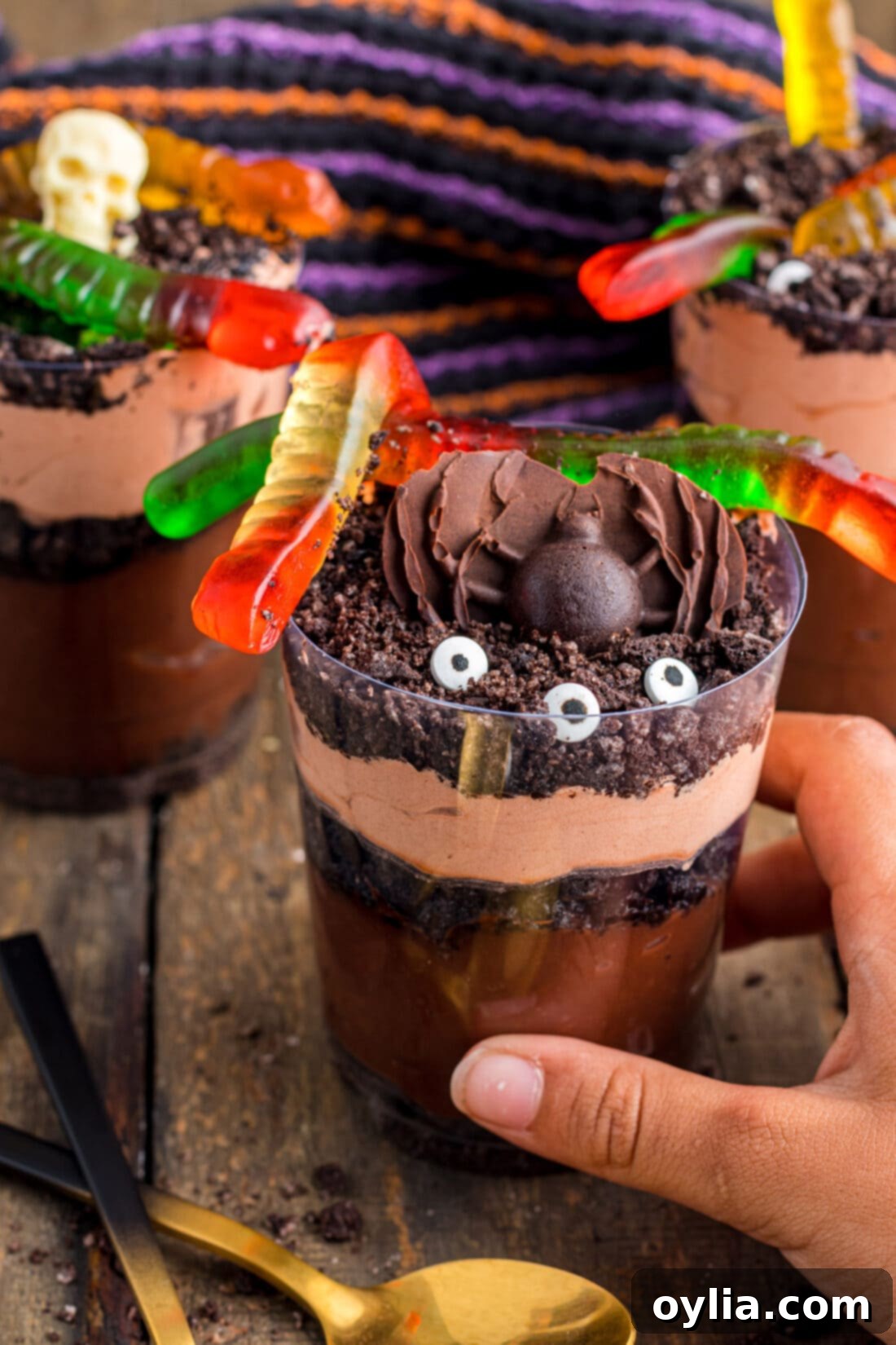 A hand reaching for a Halloween Dirt Cup, garnished with gummy worms and candy eyes.