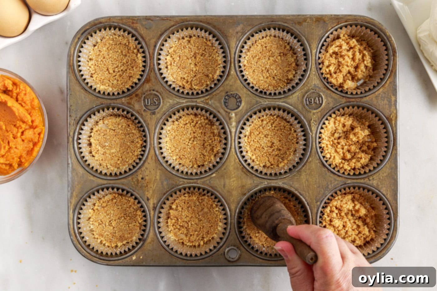 Close-up of graham cracker crust being pressed into a cupcake liner in a muffin tin.