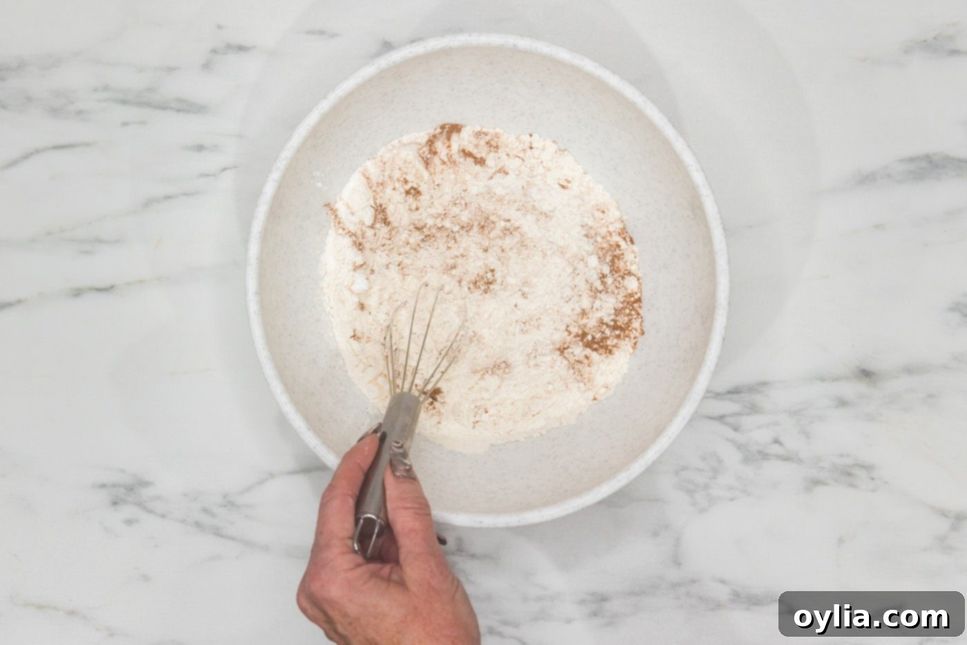Hand whisking flour, cinnamon, baking soda, and baking powder in a bowl, showing the combined dry ingredients.