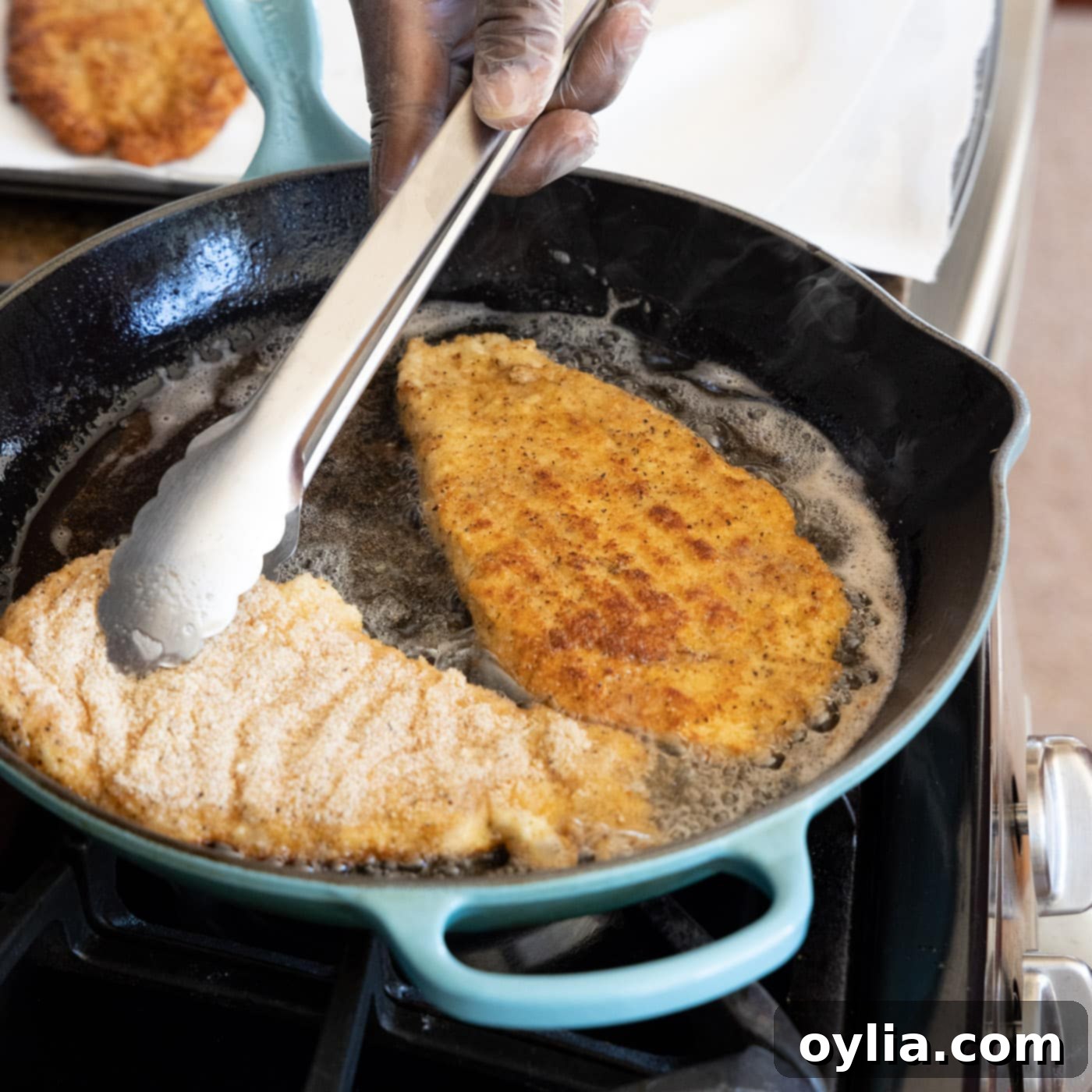 Using tongs to carefully flip a chicken Milanese cutlet in a hot skillet, revealing a perfectly golden-brown underside.