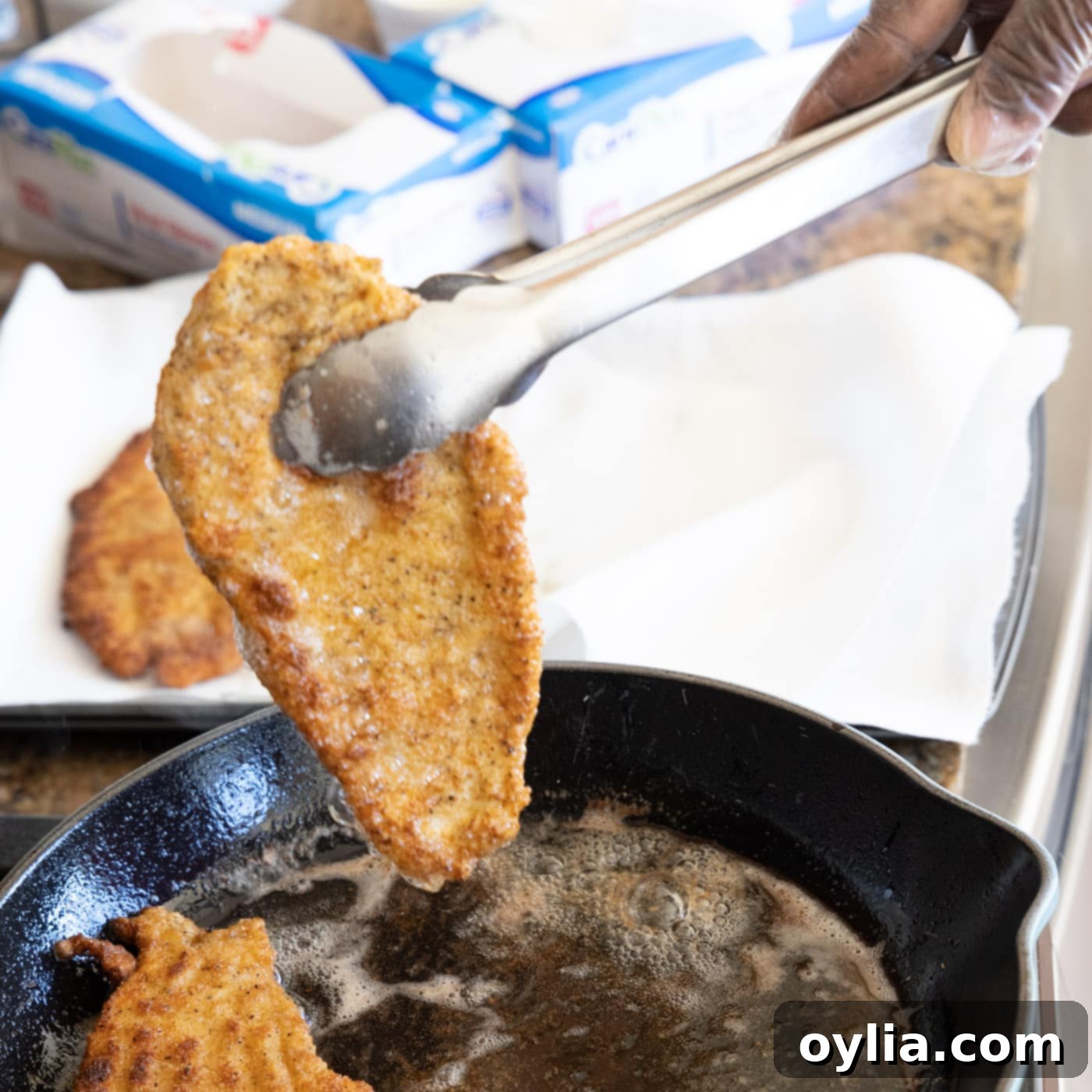 Removing a freshly fried, golden brown chicken Milanese cutlet from a skillet and placing it on a wire rack to drain.