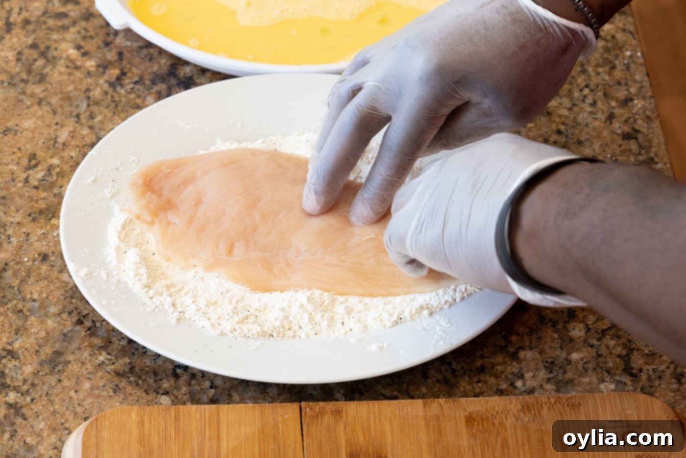 A gloved hand pressing a chicken cutlet into a plate of seasoned flour, ensuring complete coverage.