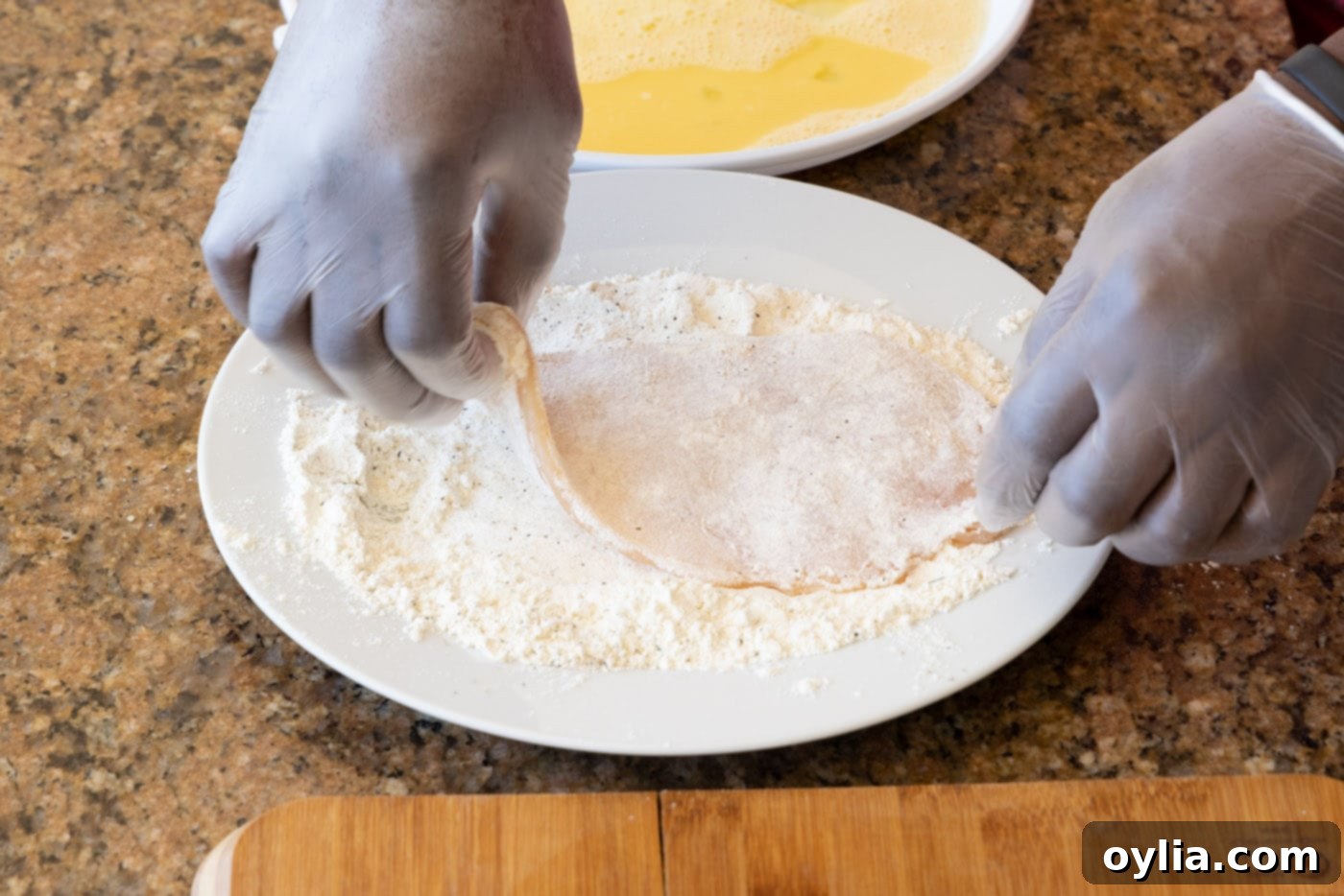 A close-up of a hand gently dredging a floured chicken cutlet, ensuring excess flour is removed for perfect breading.