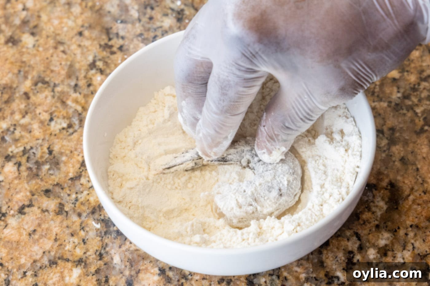 A hand dipping a raw shrimp into a bowl of all-purpose flour, ensuring it's fully coated.
