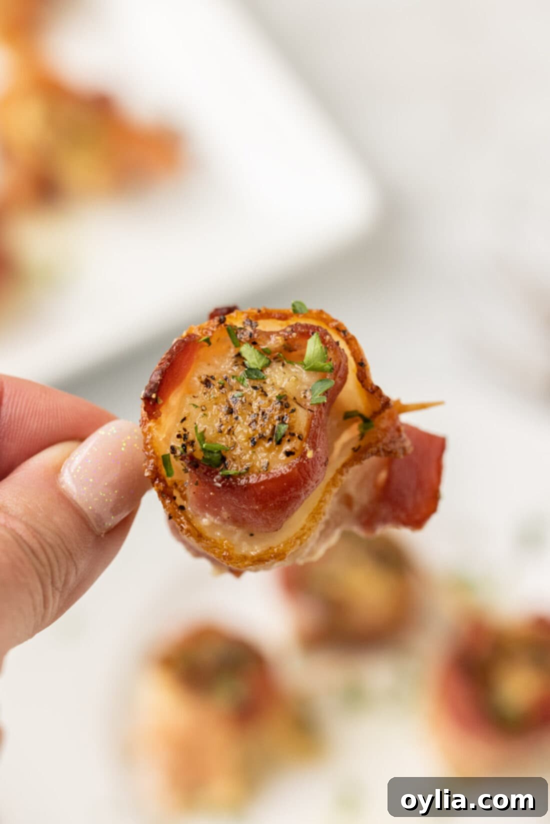 A hand holding a single perfectly cooked bacon wrapped scallop, showcasing its golden-brown exterior and juicy interior.