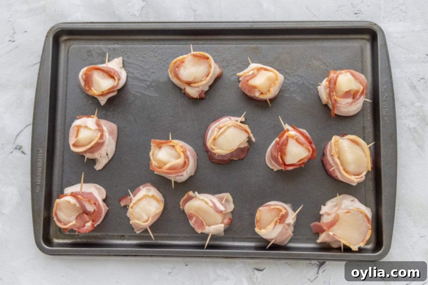 Raw sea scallops meticulously wrapped in strips of bacon and secured with toothpicks, arranged on a baking sheet before cooking.