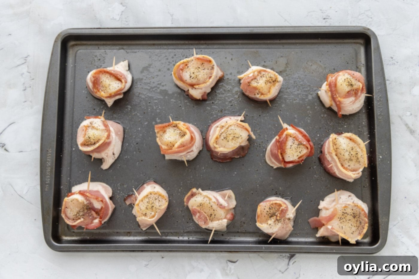 Bacon wrapped scallops seasoned with olive oil, garlic salt, and pepper, glistening on a baking sheet, ready for the broiler.