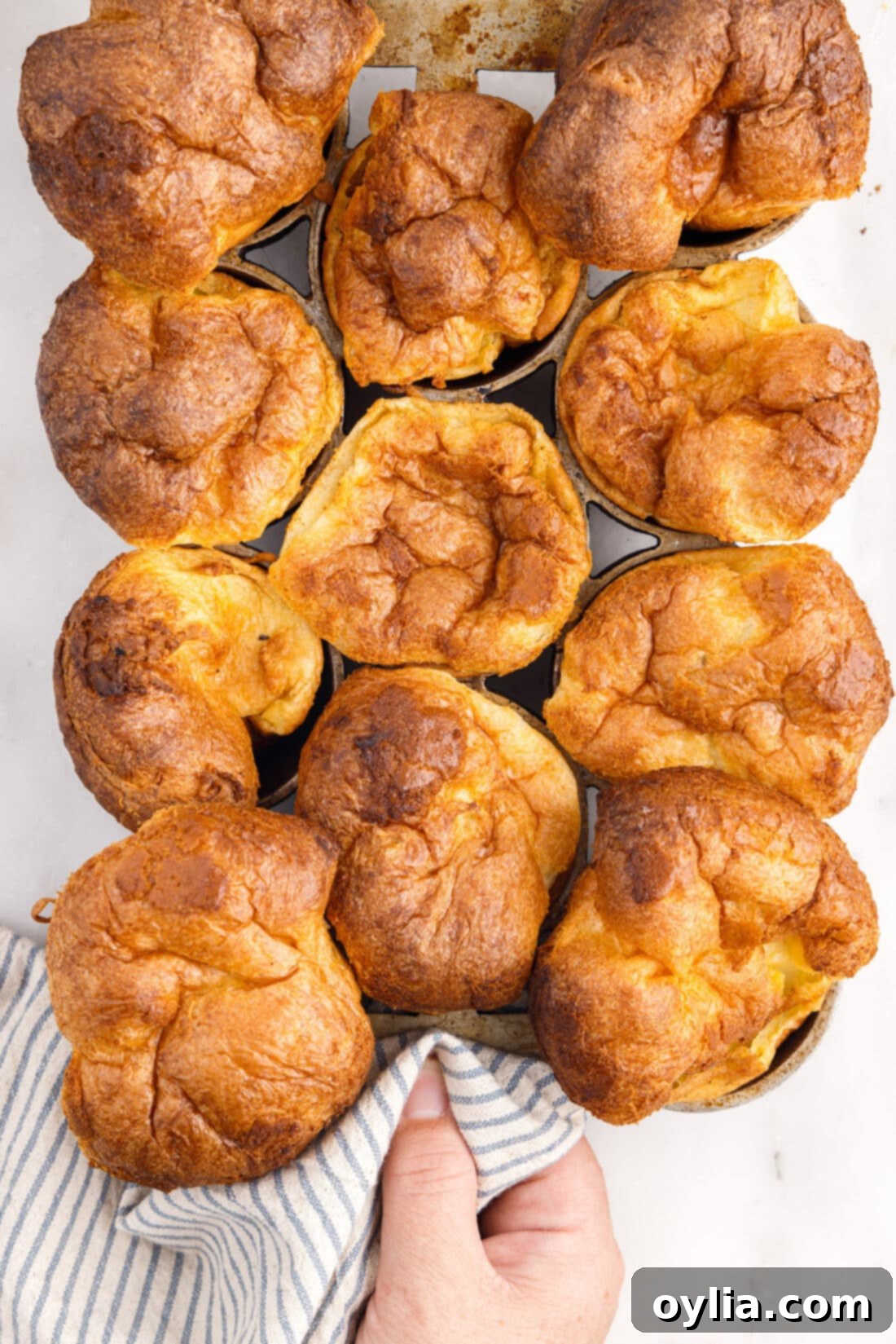 A baker carefully lifting a hot popover pan filled with tall, puffed popovers from the oven.