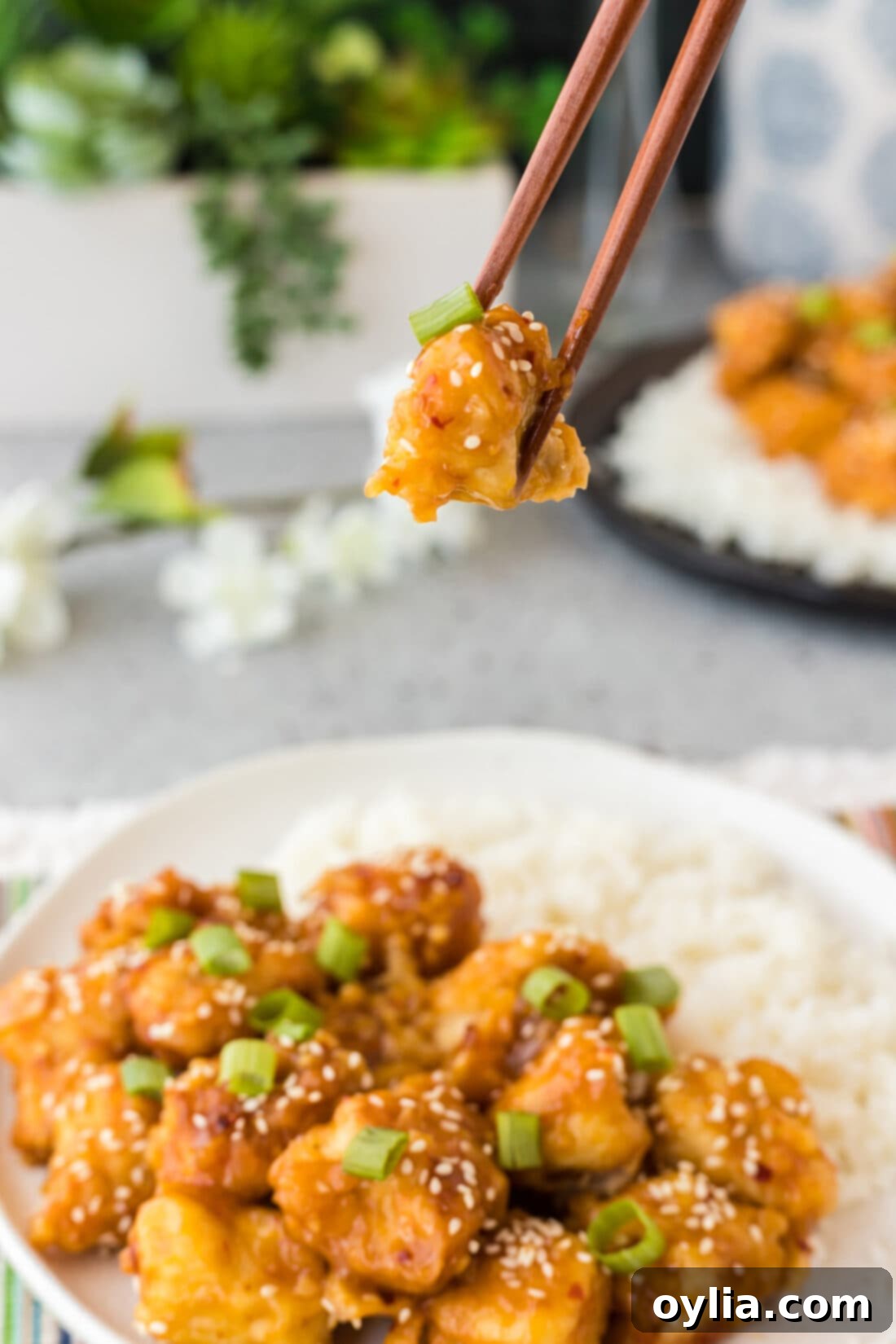 A piece of crispy orange chicken held by chopsticks, showing its sticky glaze and fluffy rice in the background