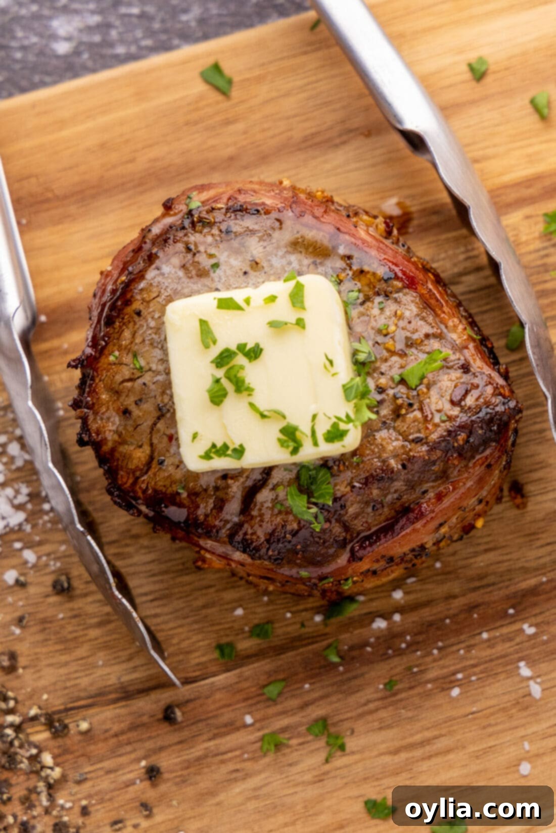 Close up of Bacon Wrapped Filet Mignon on a platter with tongs, highlighting the crispy bacon and juicy interior.