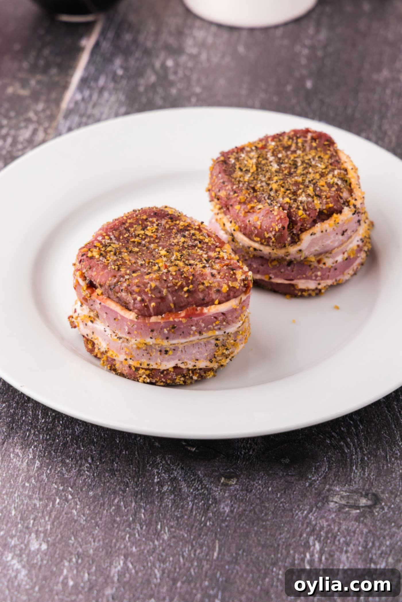 Raw filet mignon with a slice of bacon wrapped around its perimeter, secured with a toothpick, on a plate.