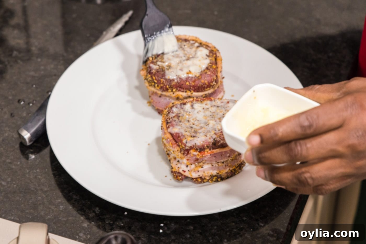 A hand brushing melted butter onto the top surface of a raw bacon-wrapped filet mignon.