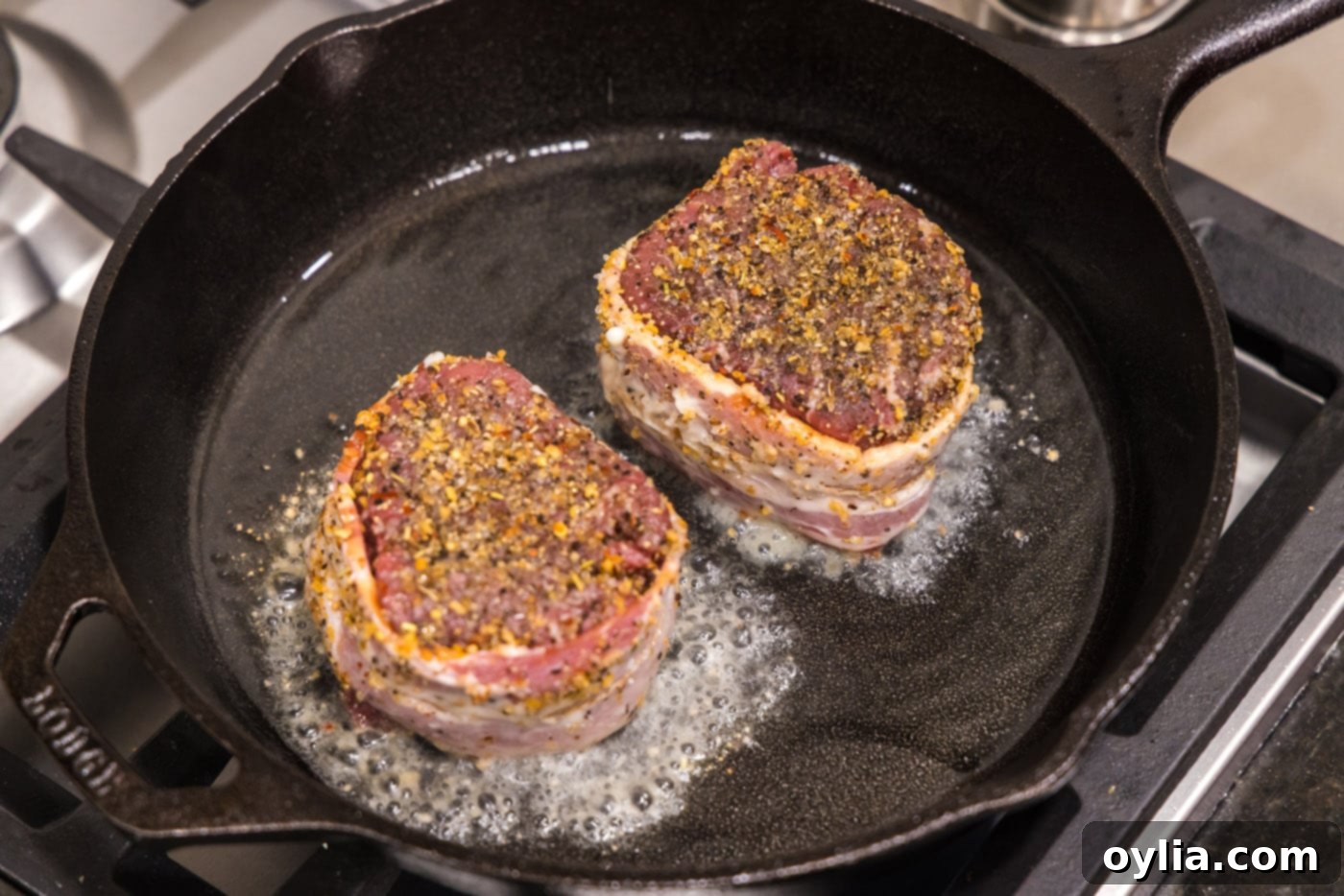 Two bacon wrapped filet mignons searing in hot oil in a skillet, butter side down.