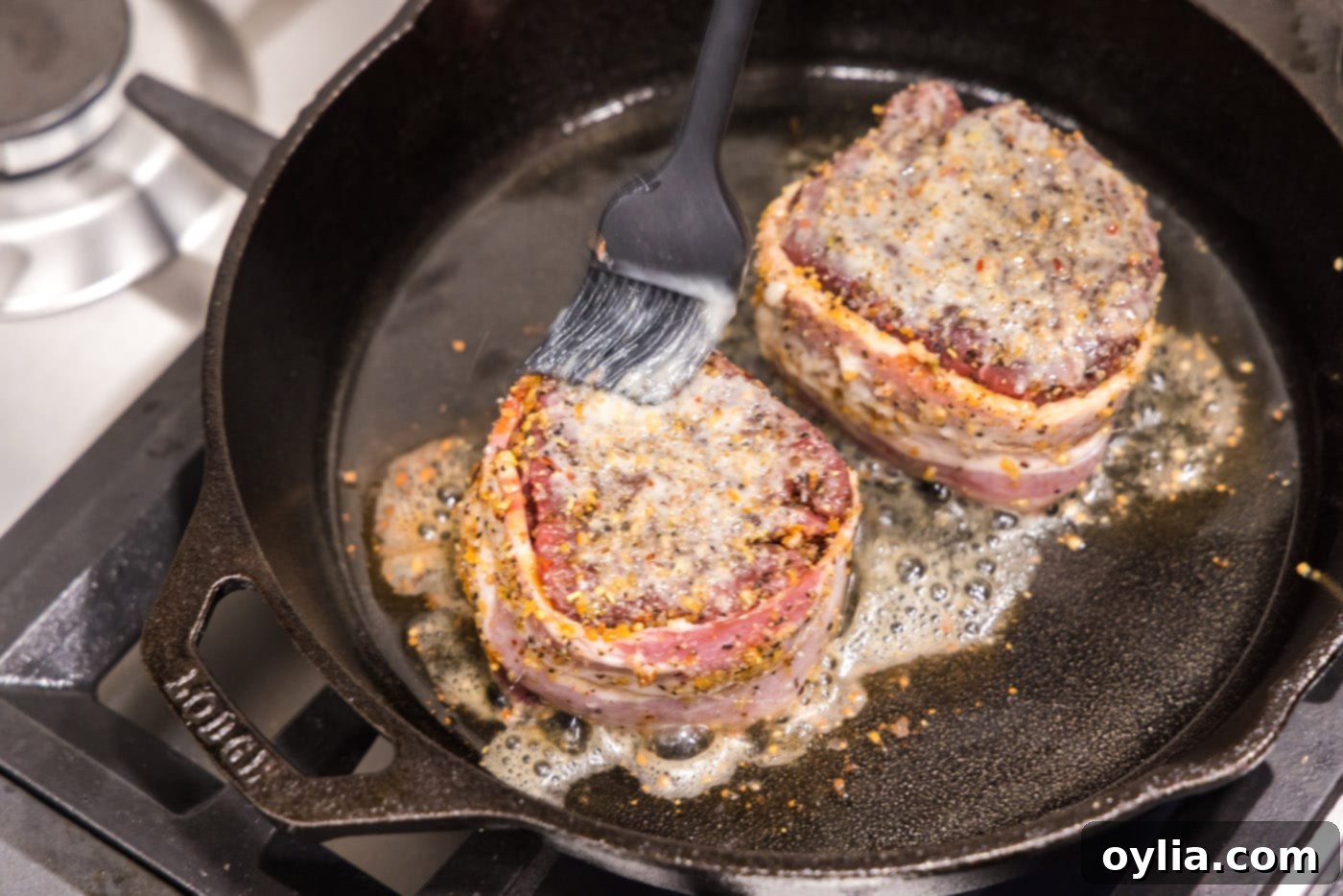 A hand brushing butter onto the top of a searing bacon-wrapped filet mignon in a skillet.