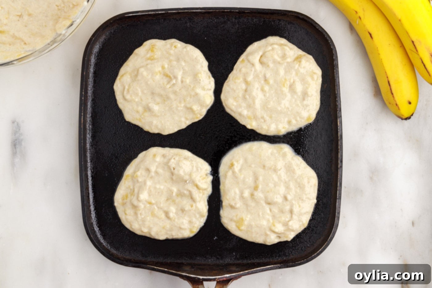 Banana pancake batter cooking on a hot griddle, forming round shapes.