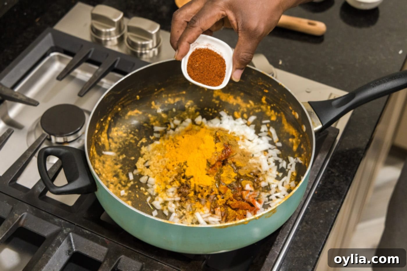 A hand adding various spices to sautéed onions in a skillet.