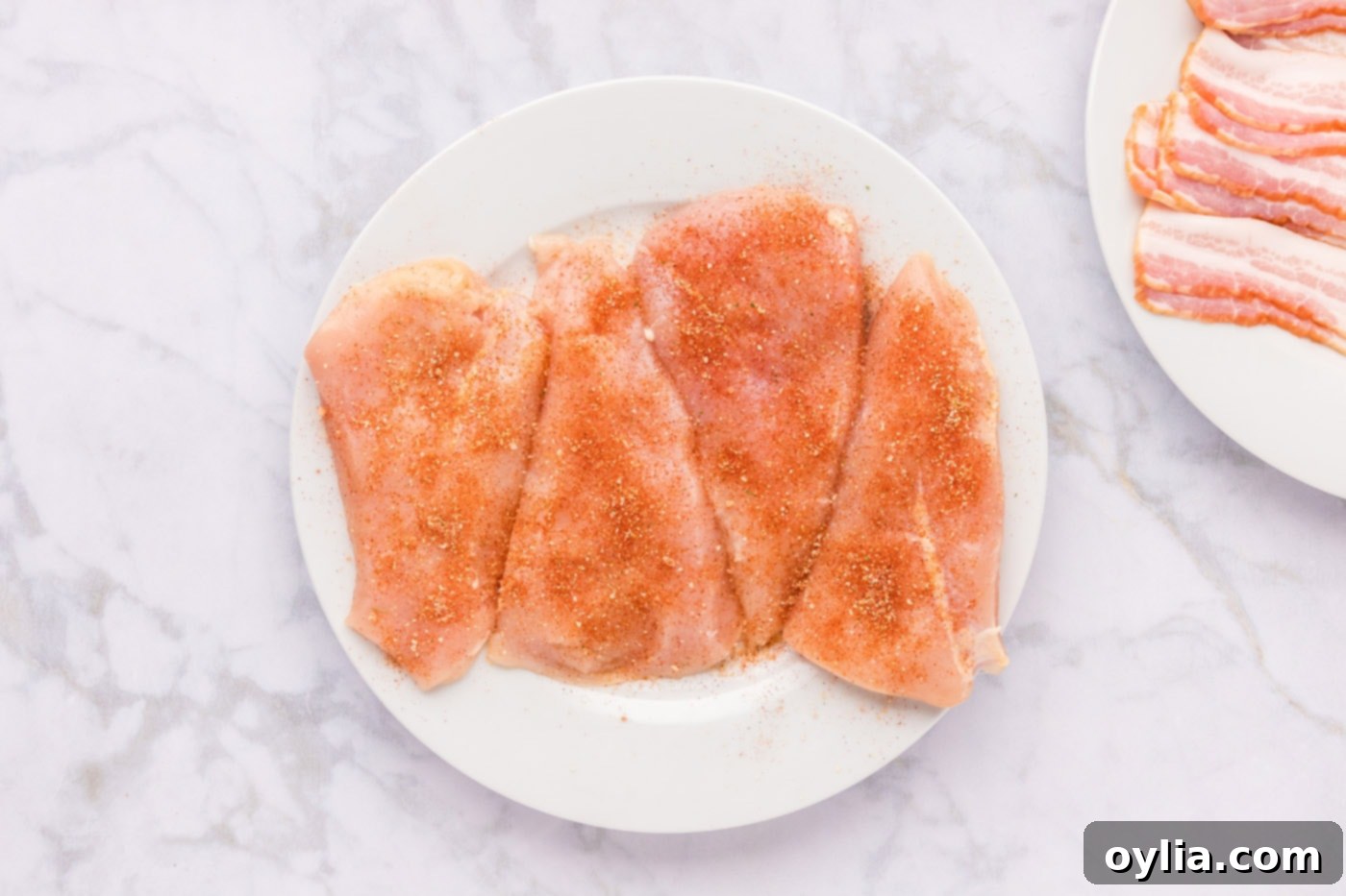 Seasoned chicken breasts on a white plate, ready for bacon wrapping