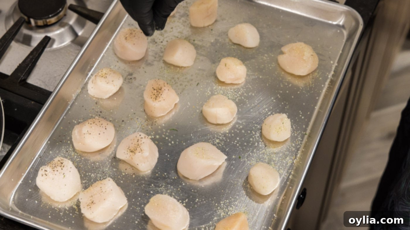 A hand sprinkling garlic salt and black pepper over raw scallops on a baking sheet