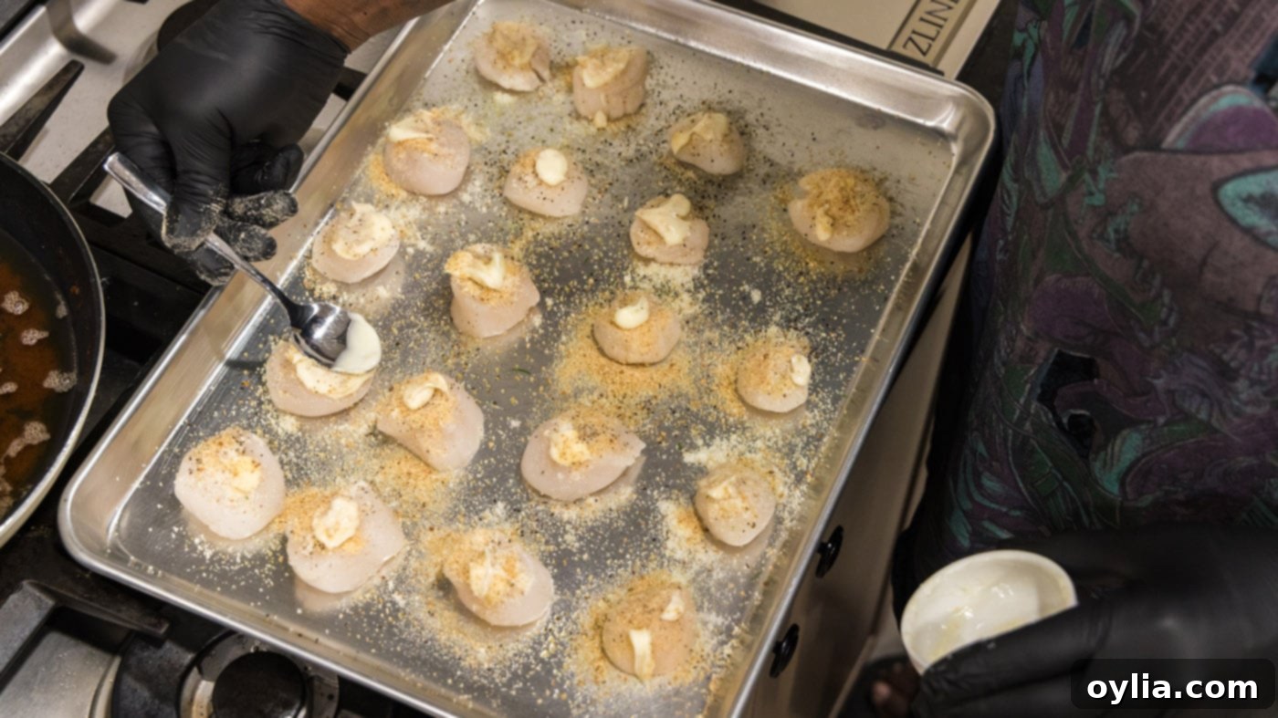 A hand gently brushing softened butter over scallops topped with breadcrumbs and parmesan