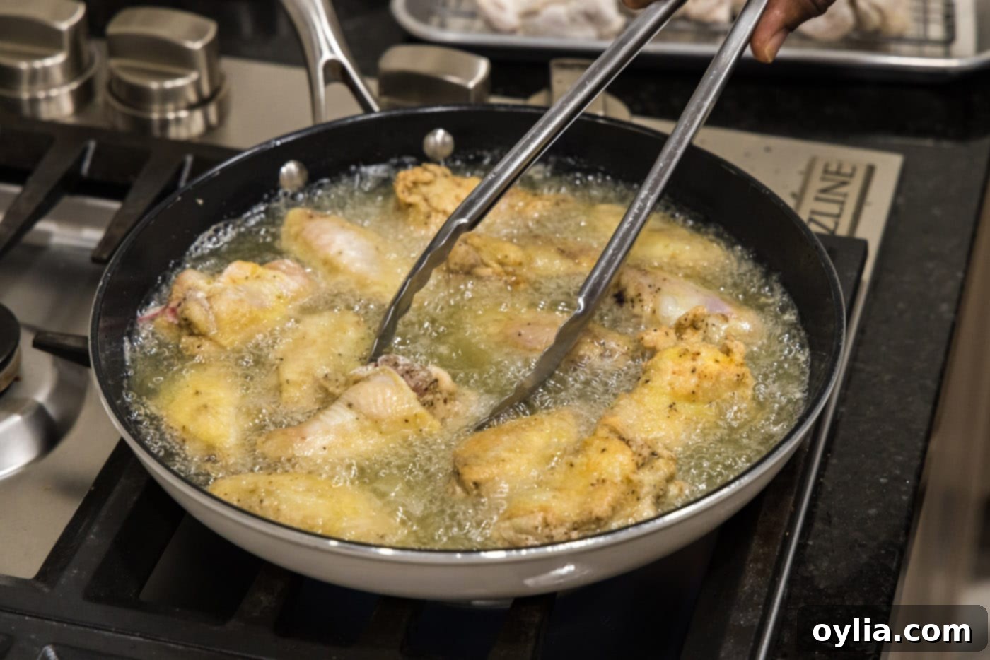 Metal tongs flipping a chicken wing in hot oil within a skillet, showing the golden-brown crust.