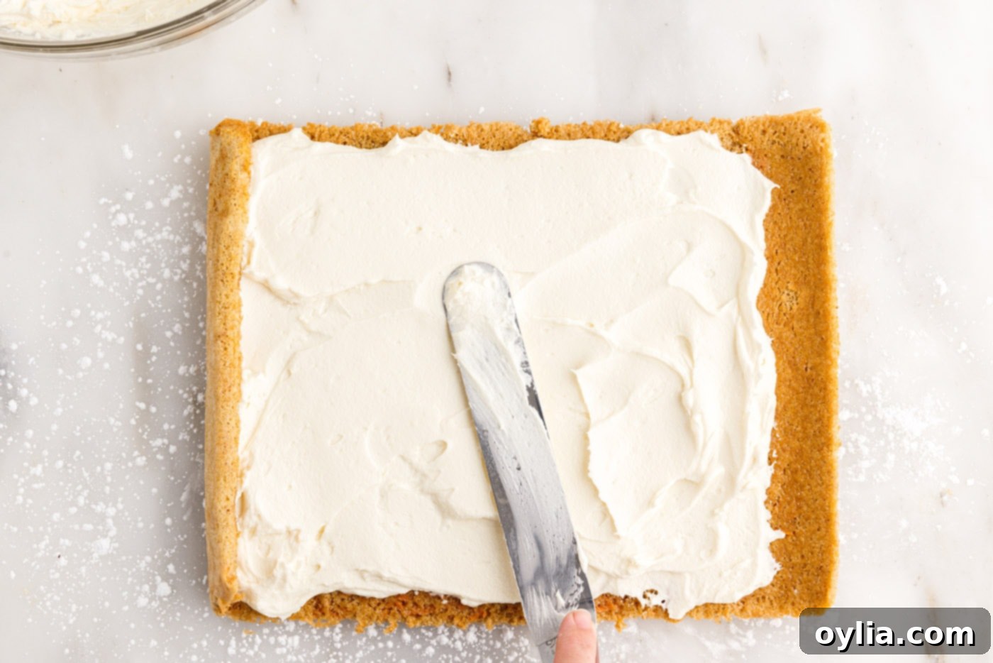 Cream cheese filling being smoothly spread across the unrolled carrot cake, leaving an edge clear