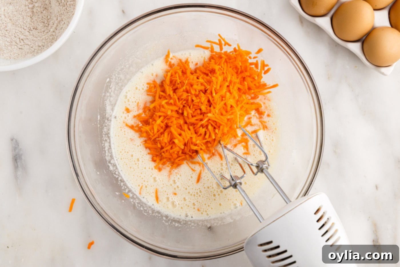 Finely grated carrots being added to the egg and sugar mixture in a bowl, with a mixer running