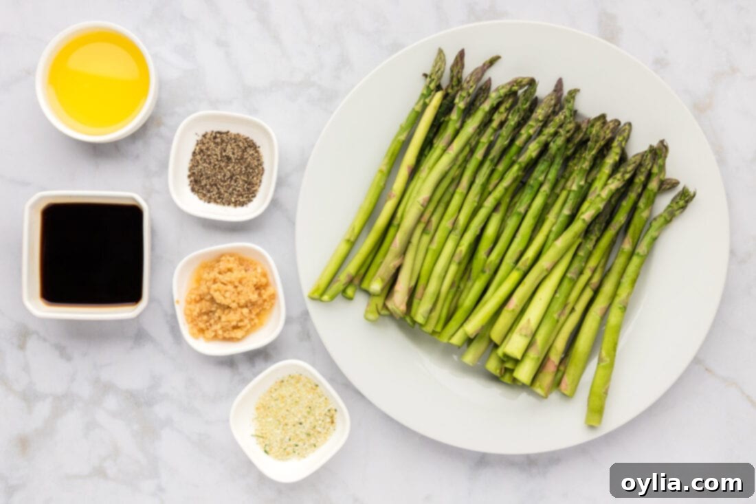 A collection of fresh ingredients laid out: a bunch of green asparagus, olive oil, a small bowl of minced garlic, garlic salt, black pepper, and a bottle of soy sauce.