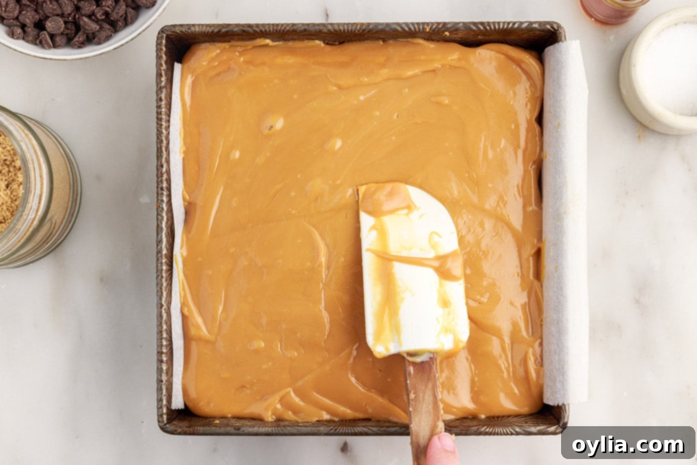 A rubber spatula smoothing warm caramel over the baked shortbread crust in the pan.