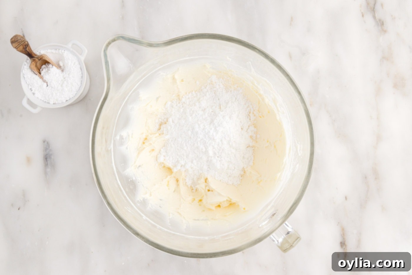 Powdered sugar is being slowly added to the cream cheese and butter mixture in a bowl, gradually transforming it into a fluffy frosting.