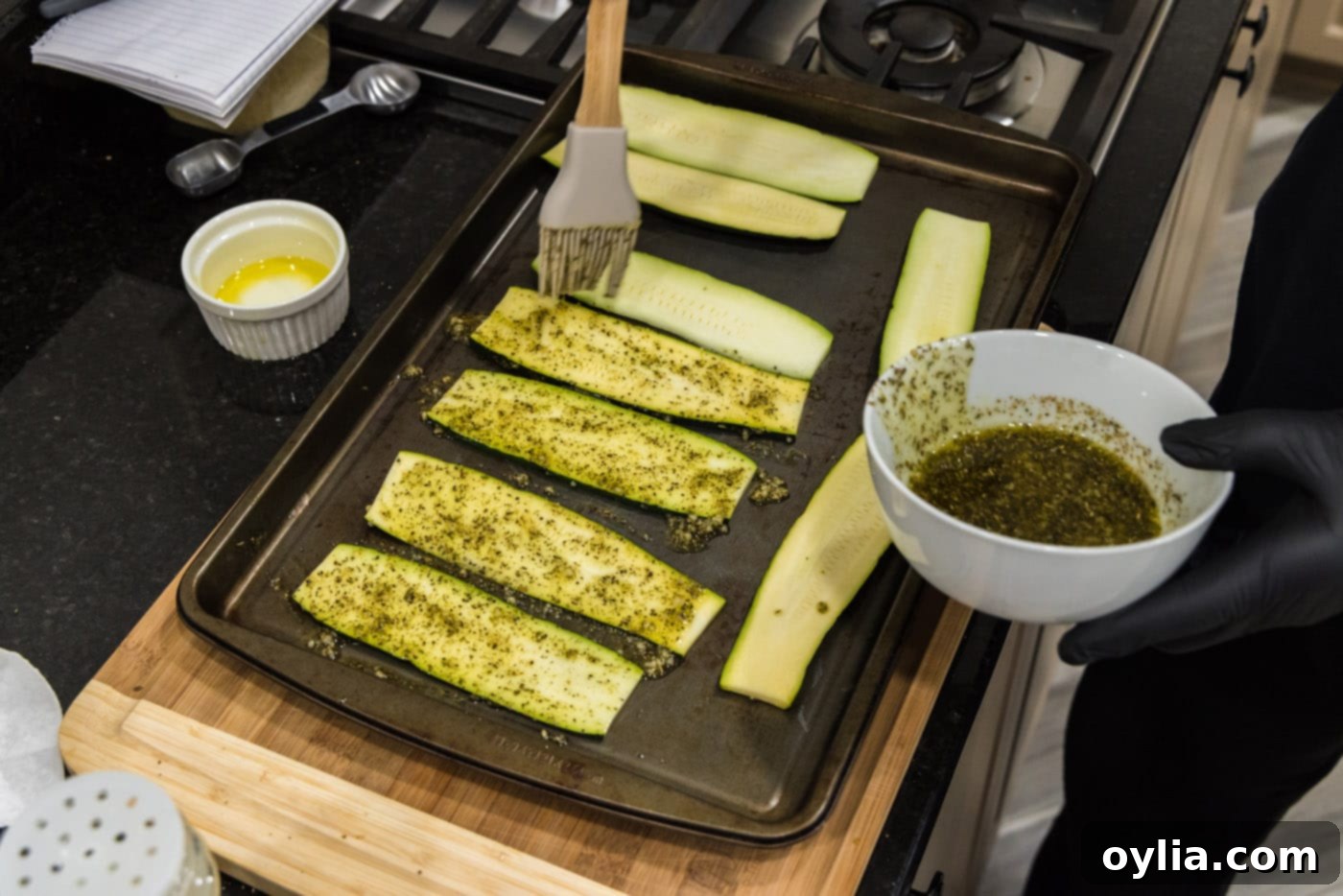 Brushing olive oil and spice mixture onto raw zucchini slices arranged on a baking sheet.