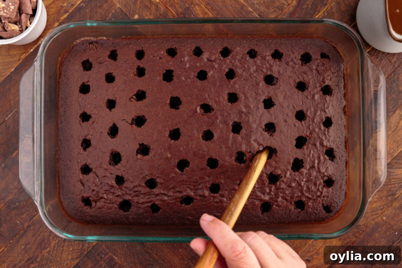 A wooden spoon handle being used to poke holes into the surface of a baked chocolate cake.