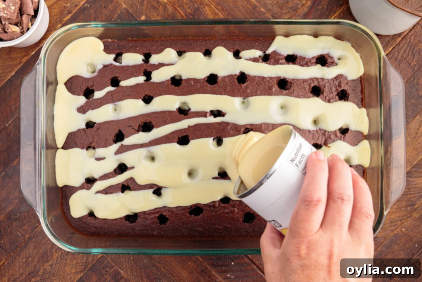 Sweetened condensed milk being poured over the poked chocolate cake.